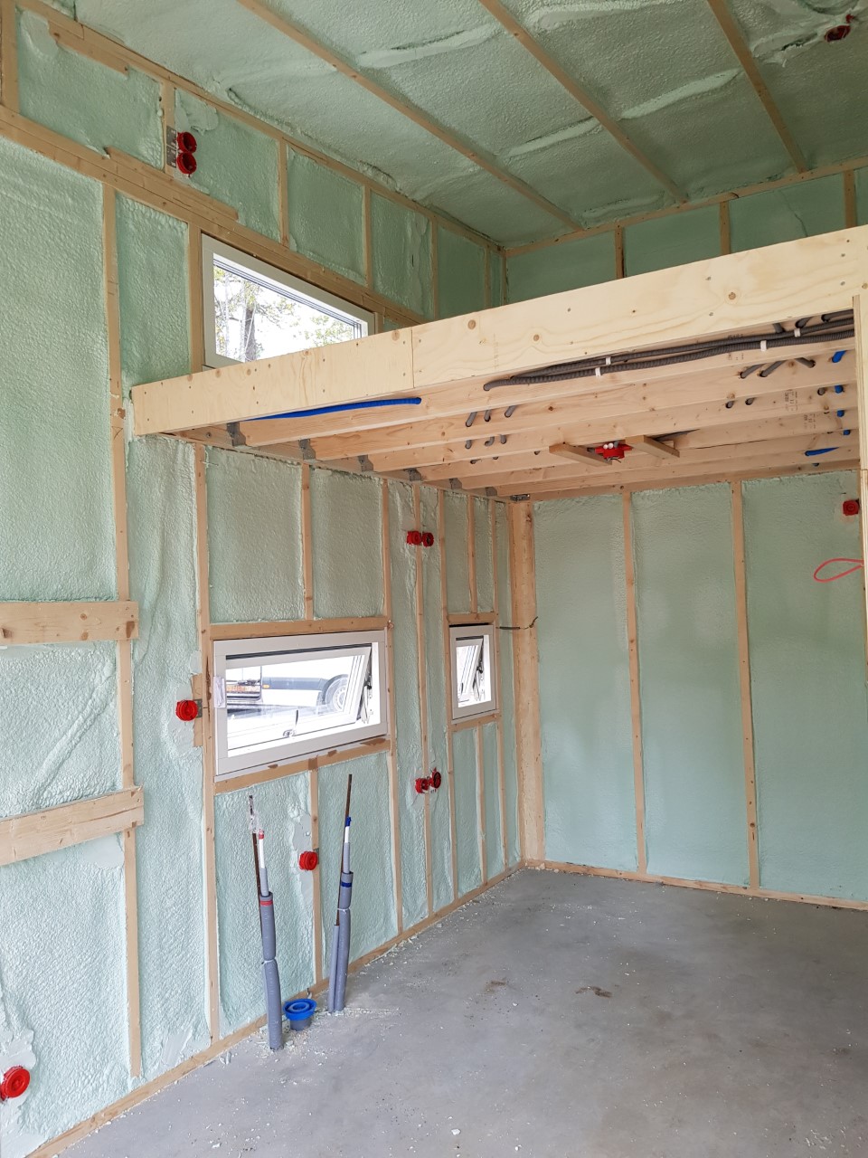 Rum under uppförande med synlig träram, sprayskumisolering på väggar och tak, synliga rör på golvet, eluttag, kablar och tre rektangulära fönster. Ett delvis loft är byggt ovanför.